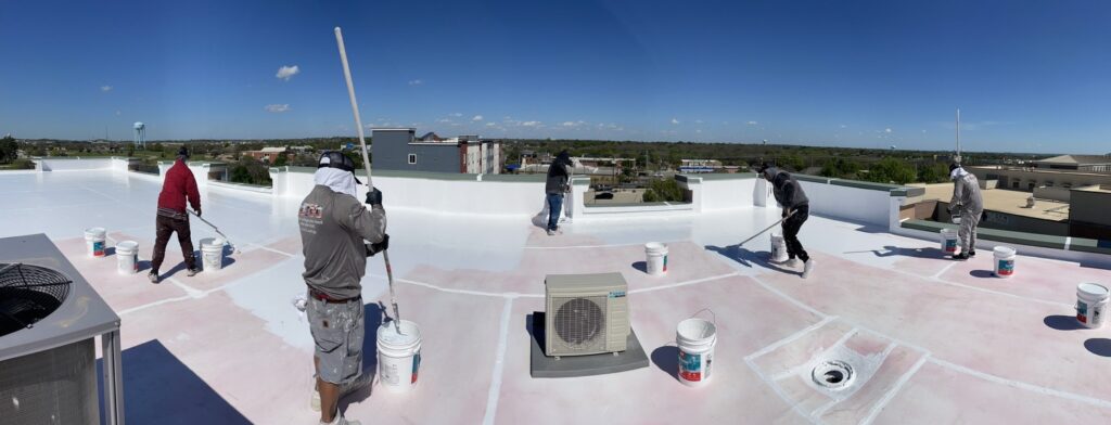 Six-man crew rolling elastomeric roof coating across a commercial flat roof with rooftop mechanical units