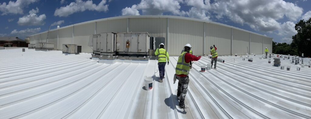 Crew applying reflective roof coating over a standing seam metal roof on a large industrial building