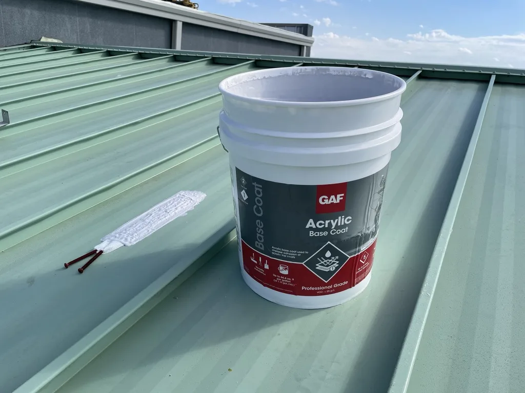 GAF Acrylic Base Coat bucket and brushes beside a prepared seam on a metal roof