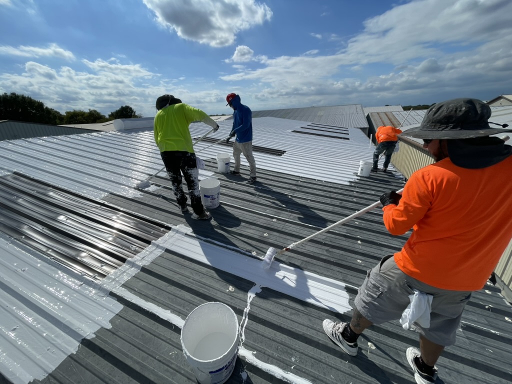 Roof coating crew applying white silicone between skylights on the McKinney R-panel roof