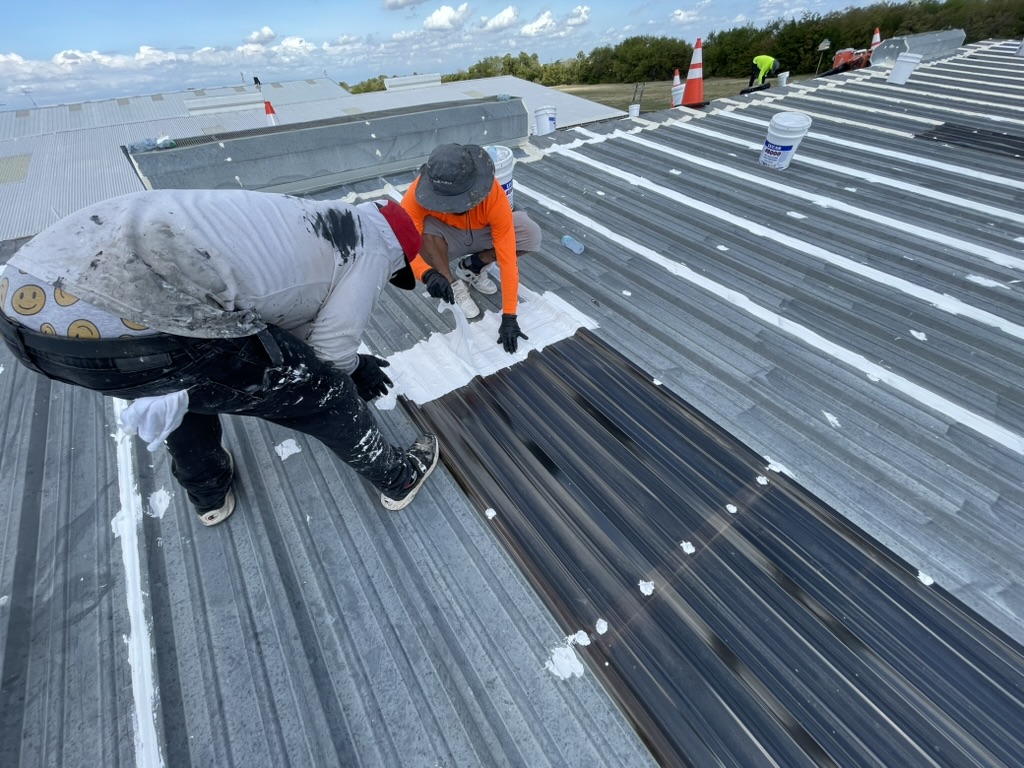 Two crew members detailing a skylight edge on the McKinney R-panel roof, with white sealant on nearby fasteners and panel seams